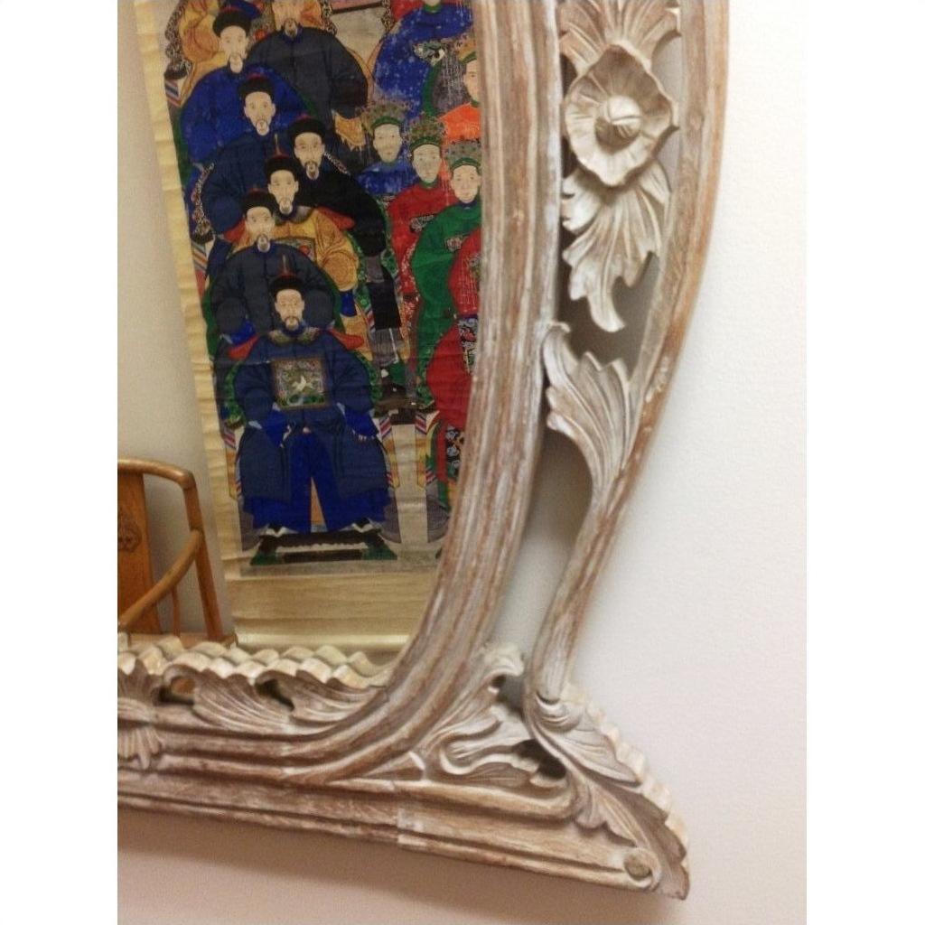 Brown carved wood mirror reflecting colorful painting of people in traditional attire on plain white wall.