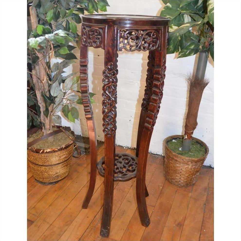 Brown carved wood plant stand with ornate details, placed on a wooden floor between two green potted plants.