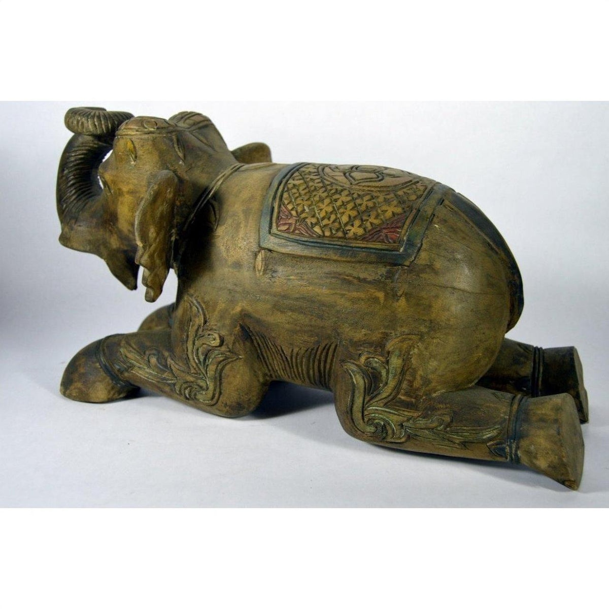 Brown wood hand-carved elephant figurine with decorated saddle, kneeling, trunk raised, close-up on neutral background.