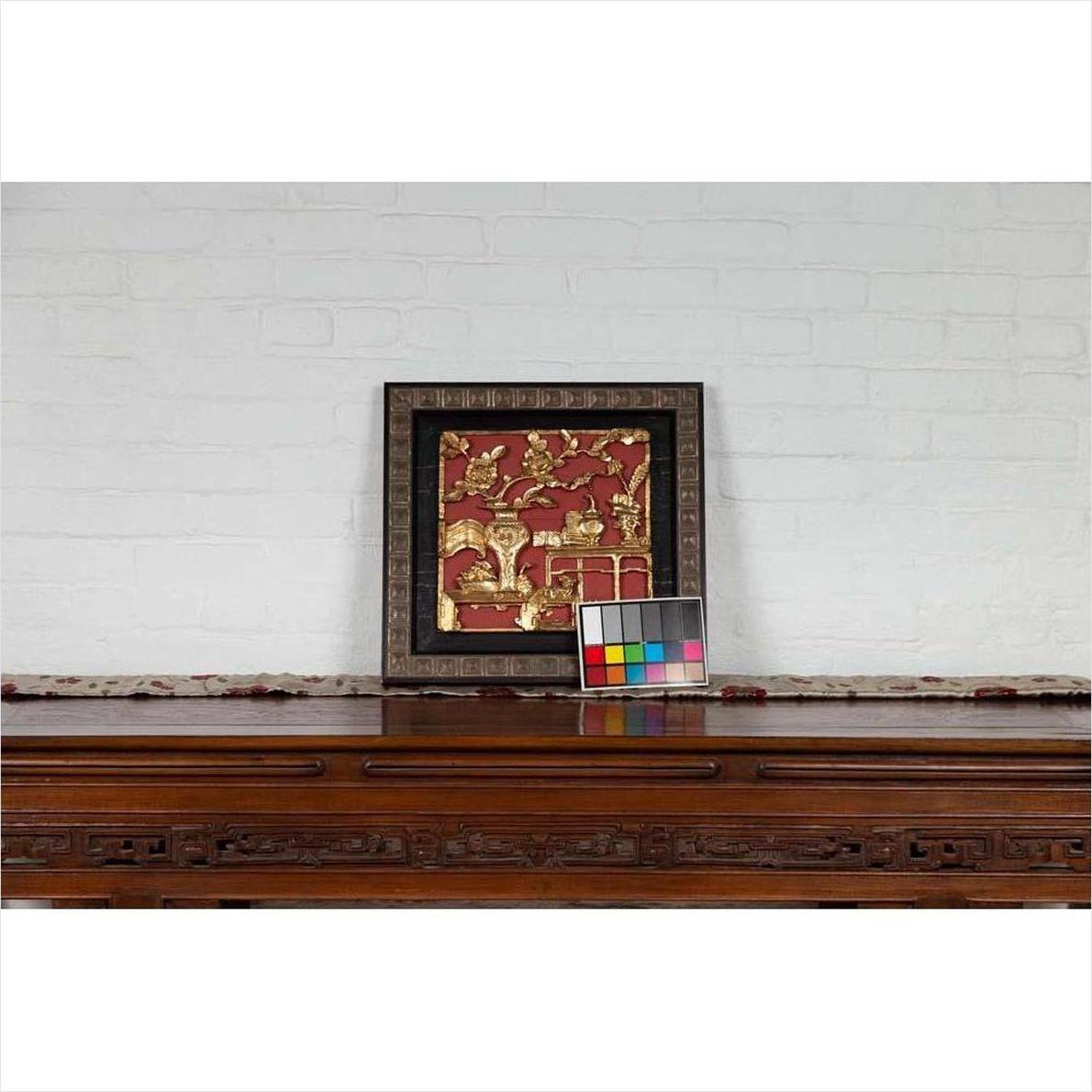 Red giltwood Chinese antique floral architectural panel on a wooden table, white brick wall background.