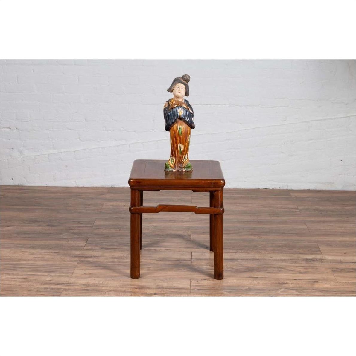 Ceramic figurine in traditional dress on brown Ming style side table, white brick wall background.