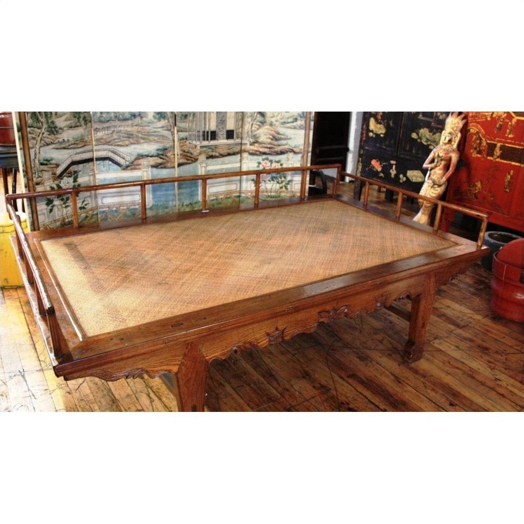 Brown wooden Chinese daybed with woven rattan surface on wood floor, decorative panels in the background.
