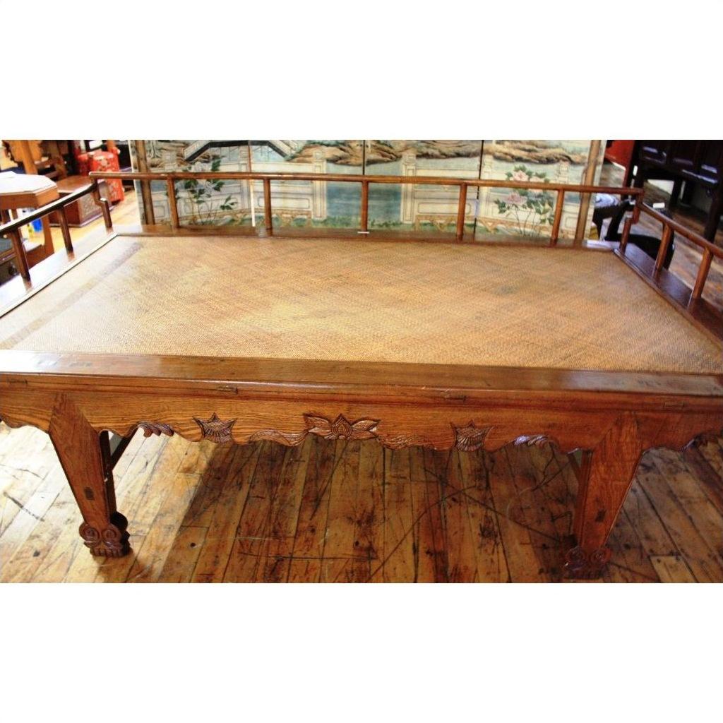 Brown wood Chinese daybed with woven rattan surface and carved legs, placed on a wooden floor.