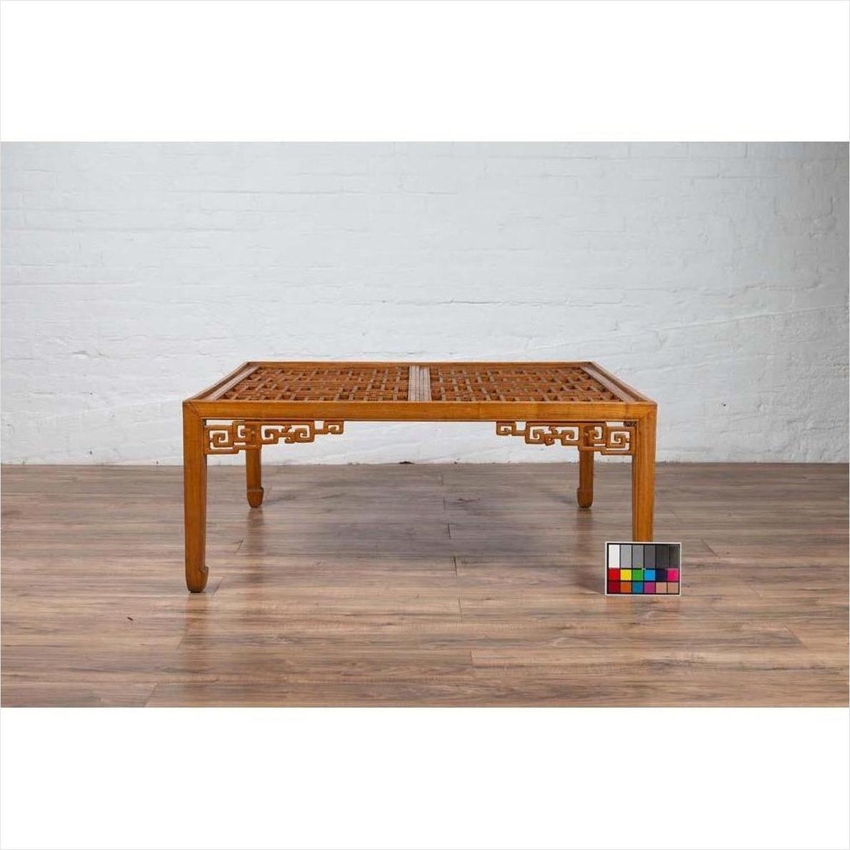 Brown elmwood square coffee table with open fretwork and horse-hoof legs on wood floor by white brick wall indoors.