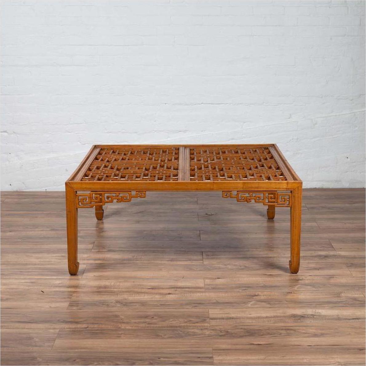 Brown elmwood square coffee table with open fretwork and horse-hoof legs on wooden floor by white brick wall indoors.