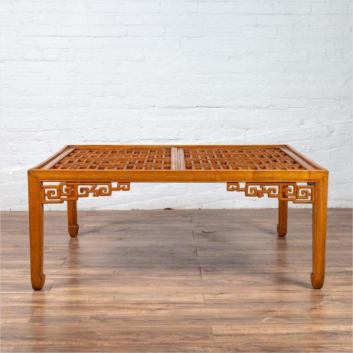 Brown elmwood square coffee table with open fretwork and horse-hoof legs on wooden floor by white brick wall indoors.