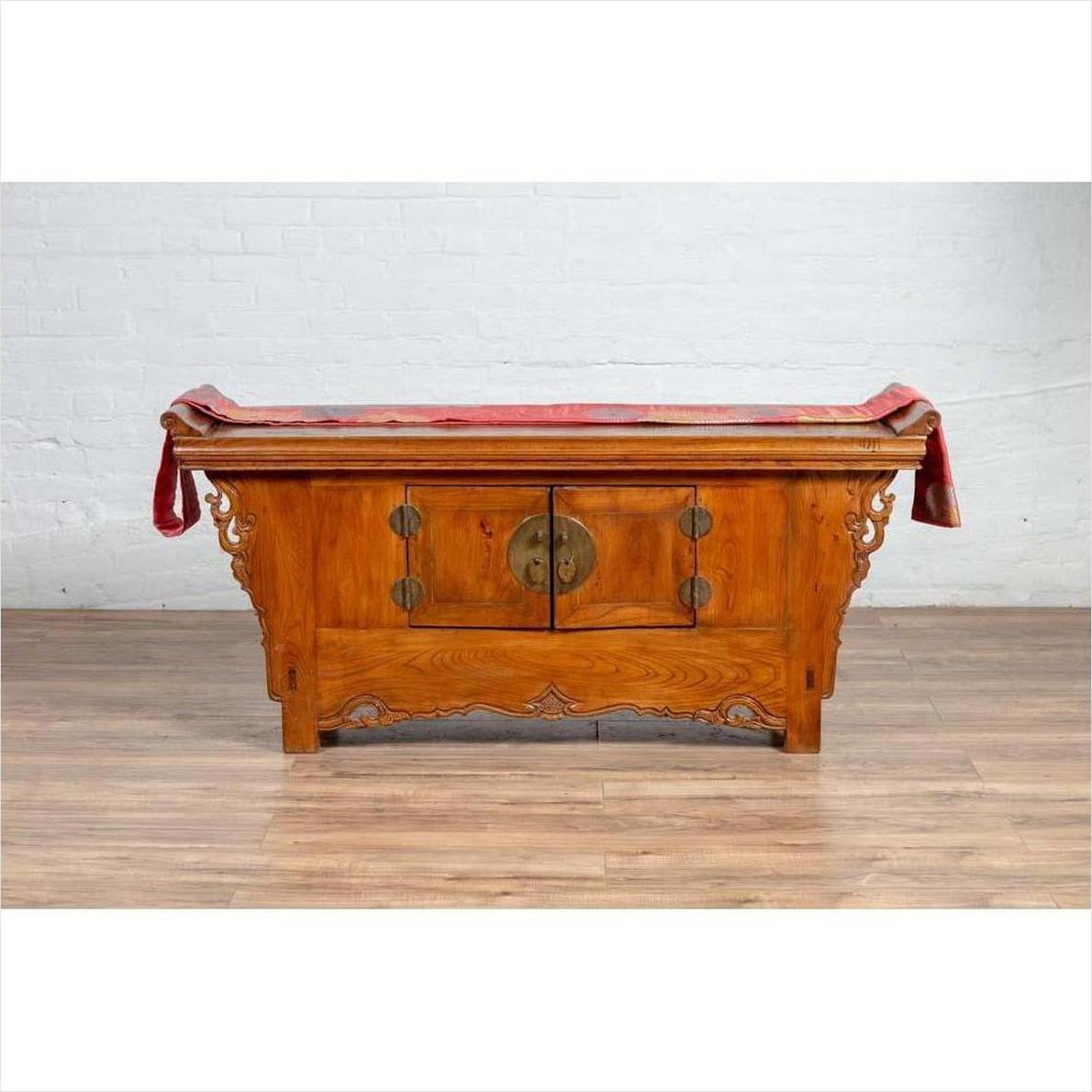 Brown elmwood two-door low kang cabinet with everted flanges, scroll details, round metal handles, red cloth on top.