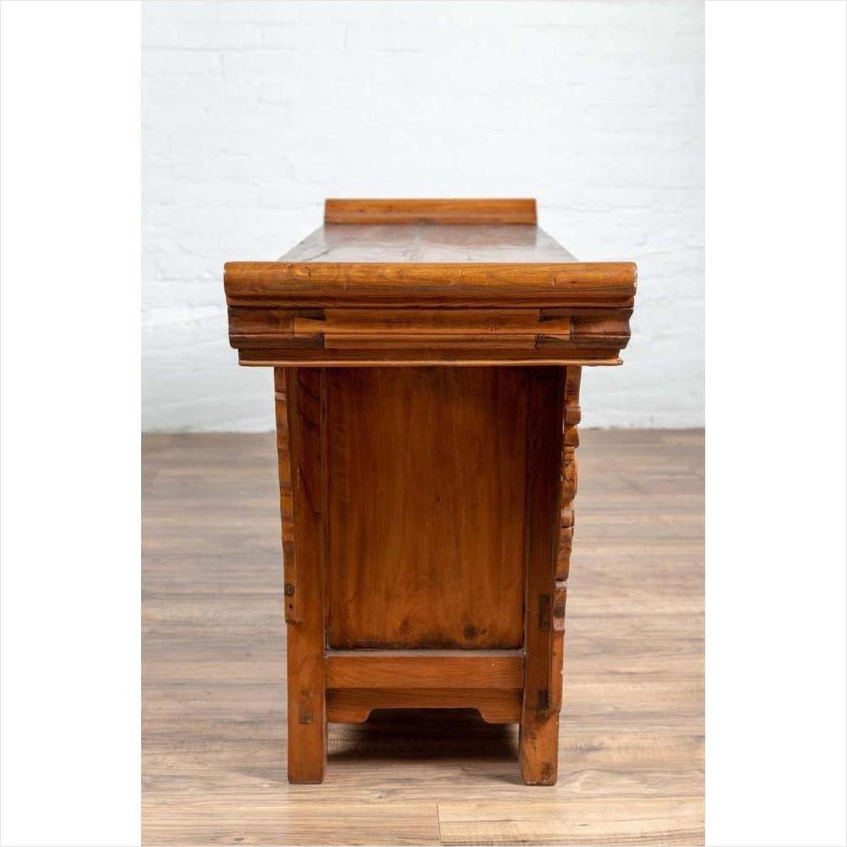 Brown elmwood two-door low cabinet with everted flanges and scrolls, side view, set against a white brick wall.