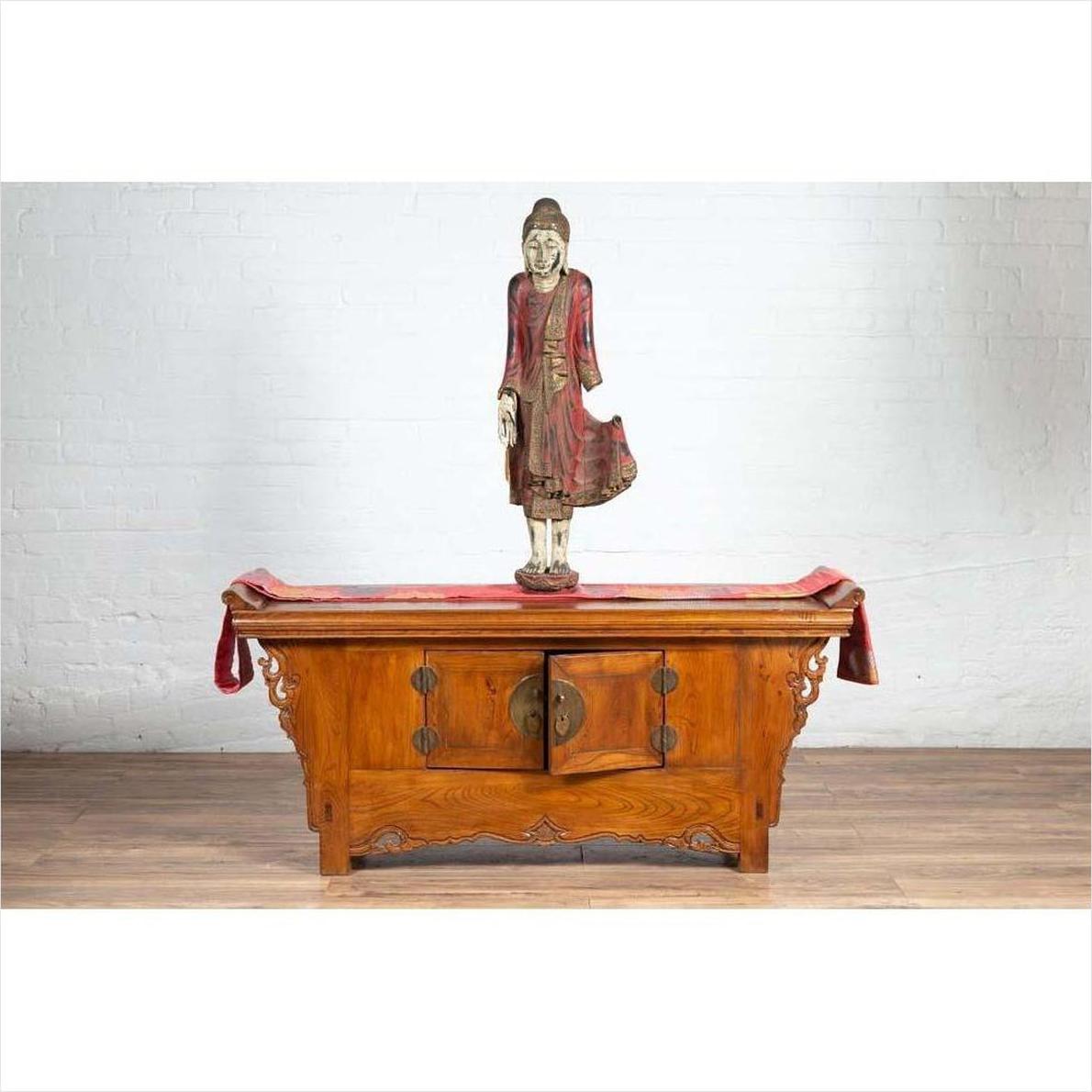 Brown wood two-door low cabinet beside wood table with red cloth and standing Buddha, on wooden floor indoors.