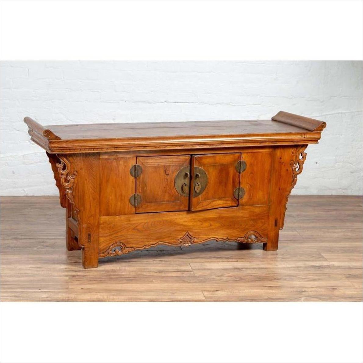 Brown elmwood low cabinet with two doors, everted flanges and scrolls, against white brick wall on wood floor.