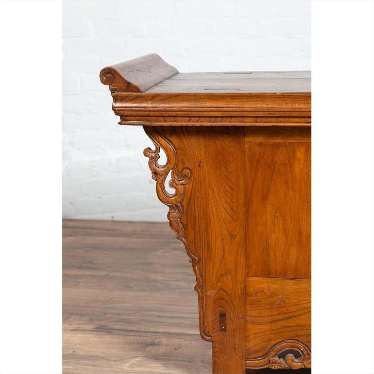 Brown elmwood low cabinet with everted flanges and scrolls, indoors against a white brick wall and wooden floor.