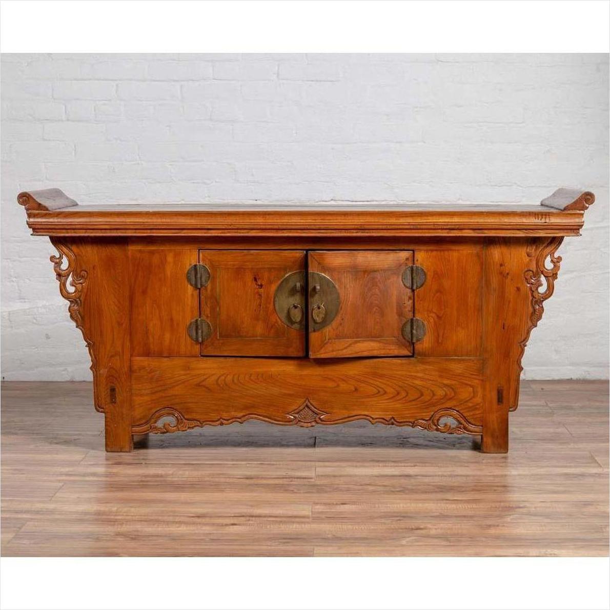 Brown elmwood two-door low cabinet with everted flanges and scrolls, indoors against a white brick wall.