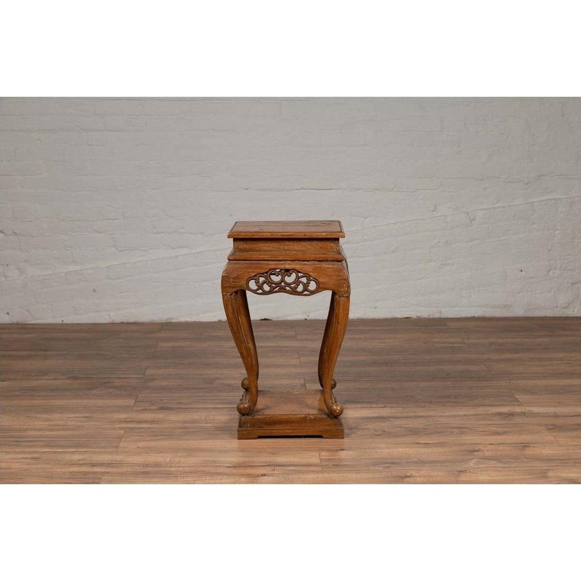 Brown wooden Ming style incense stand with cabriole legs and carved apron, placed next to a white brick wall indoors.