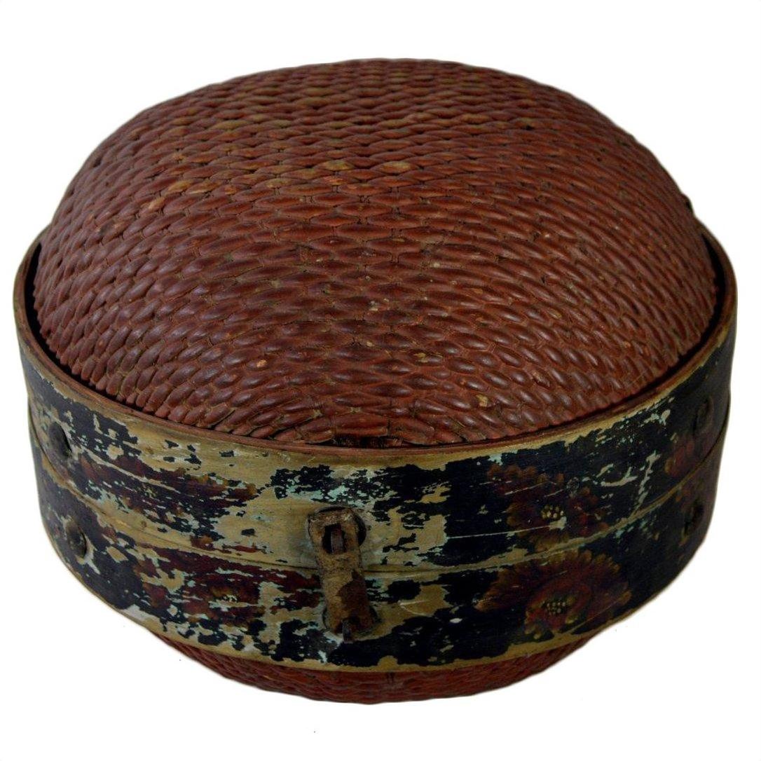 Close-up of round brown rattan Chinese storage basket with domed lid, black weathered floral band, and small lock.