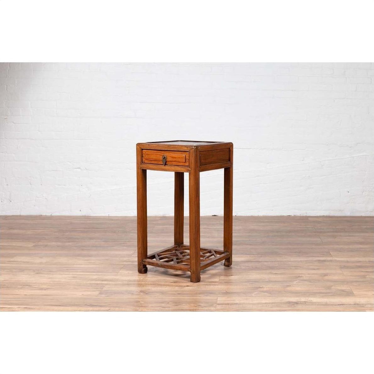 Brown elmwood lamp table with single drawer and cracked ice shelf on wooden floor next to white brick wall.