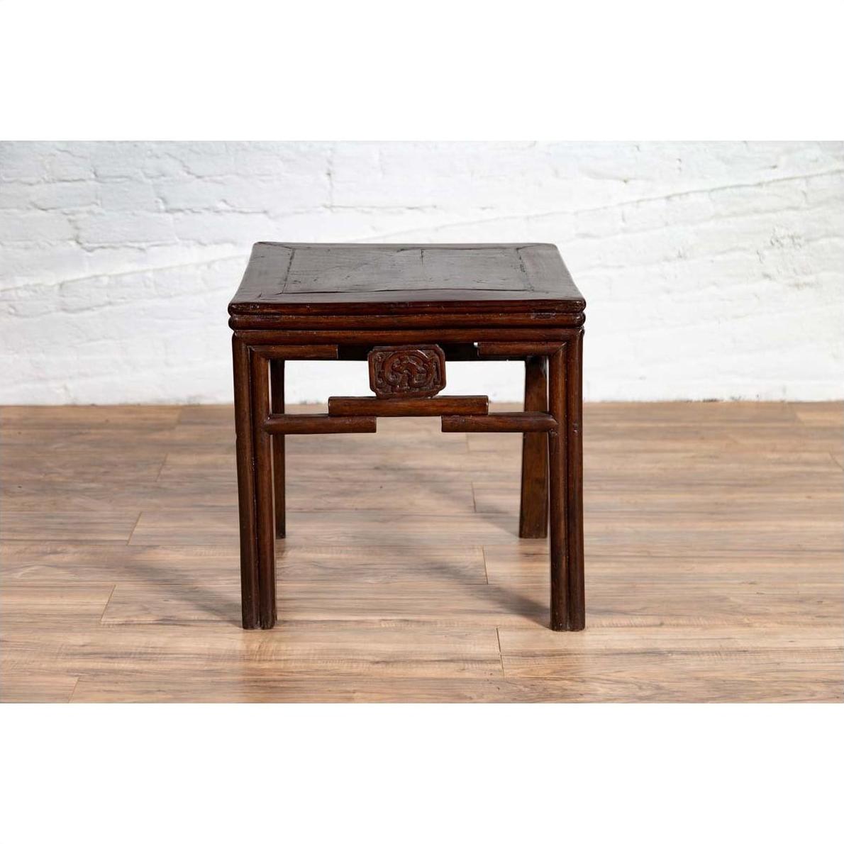 Dark wood Qing style side table with carved medallions on a wood floor next to a white brick wall.