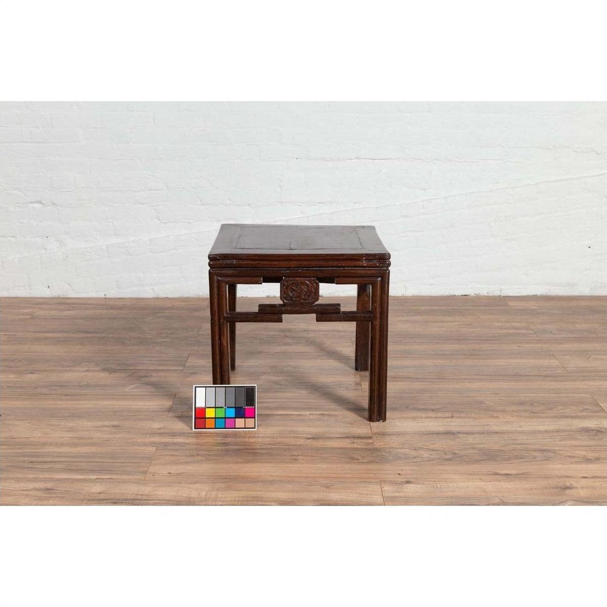 Dark wood Qing style side table with carved medallions, on wood floor next to white brick wall indoors.