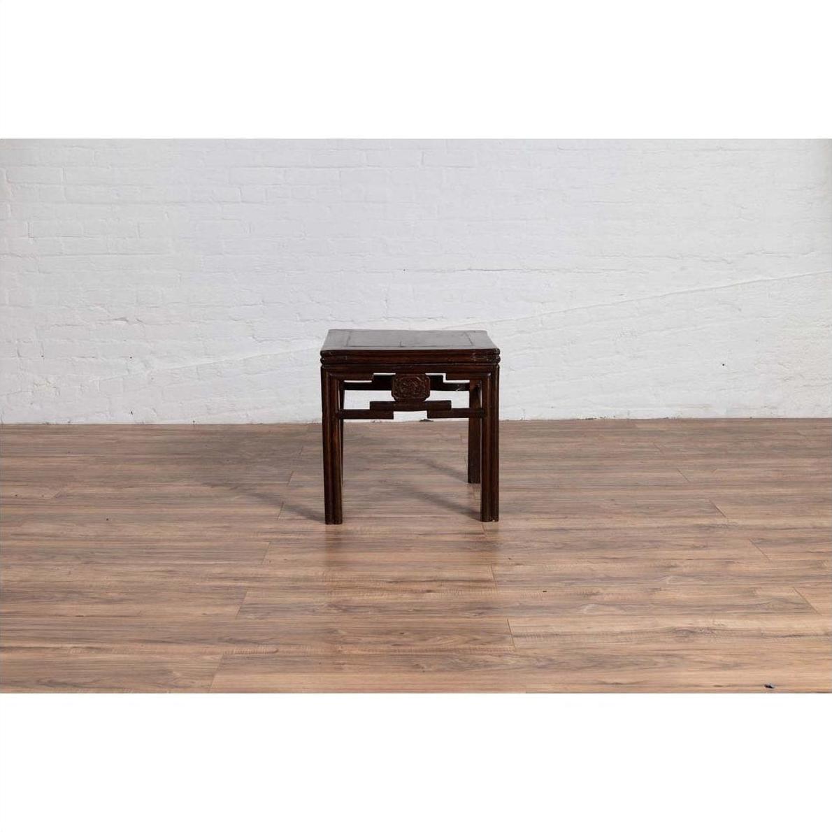 Dark wood Chinese side table with carved medallions, vintage Qing style, on wood floor by white brick wall indoors.