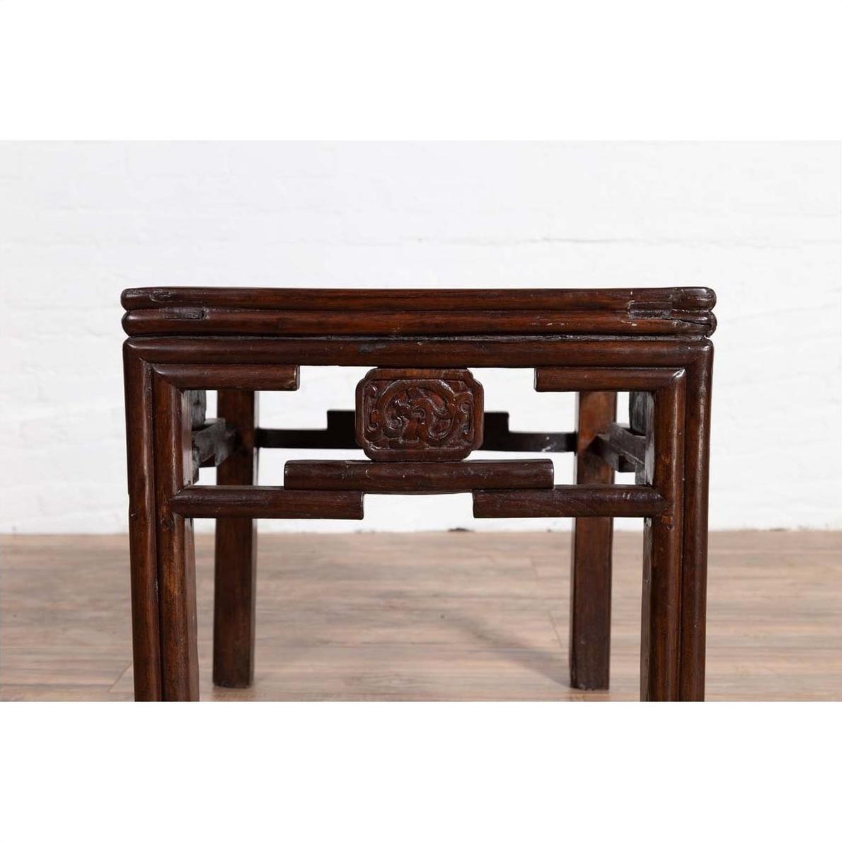 Dark wood Qing style side table with carved medallions, on wood floor against white brick wall.