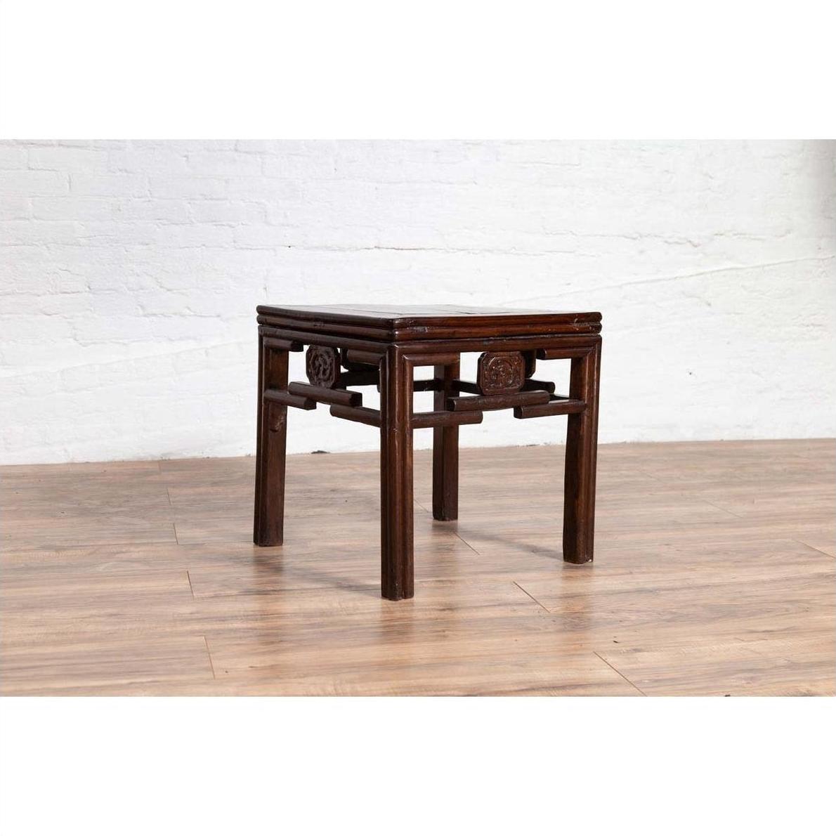 Dark wood Chinese Qing style side table with carved medallions, on wooden floor against white brick wall indoors.