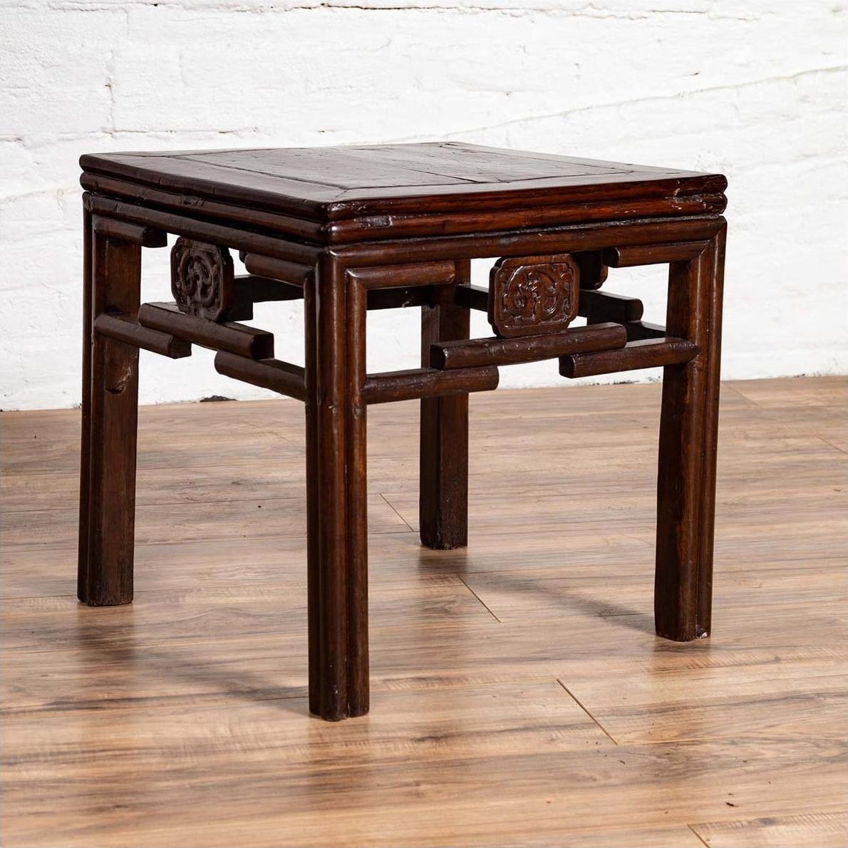 Dark wood Chinese Qing style side table with carved medallions on wooden floor next to white brick wall indoors.