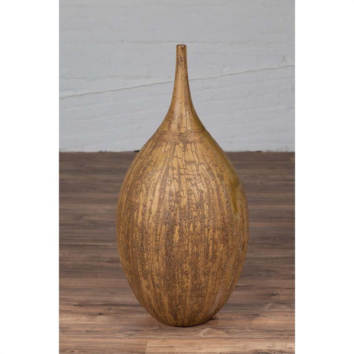 Mustard ceramic vase with tapered spout on wooden floor against white brick wall.