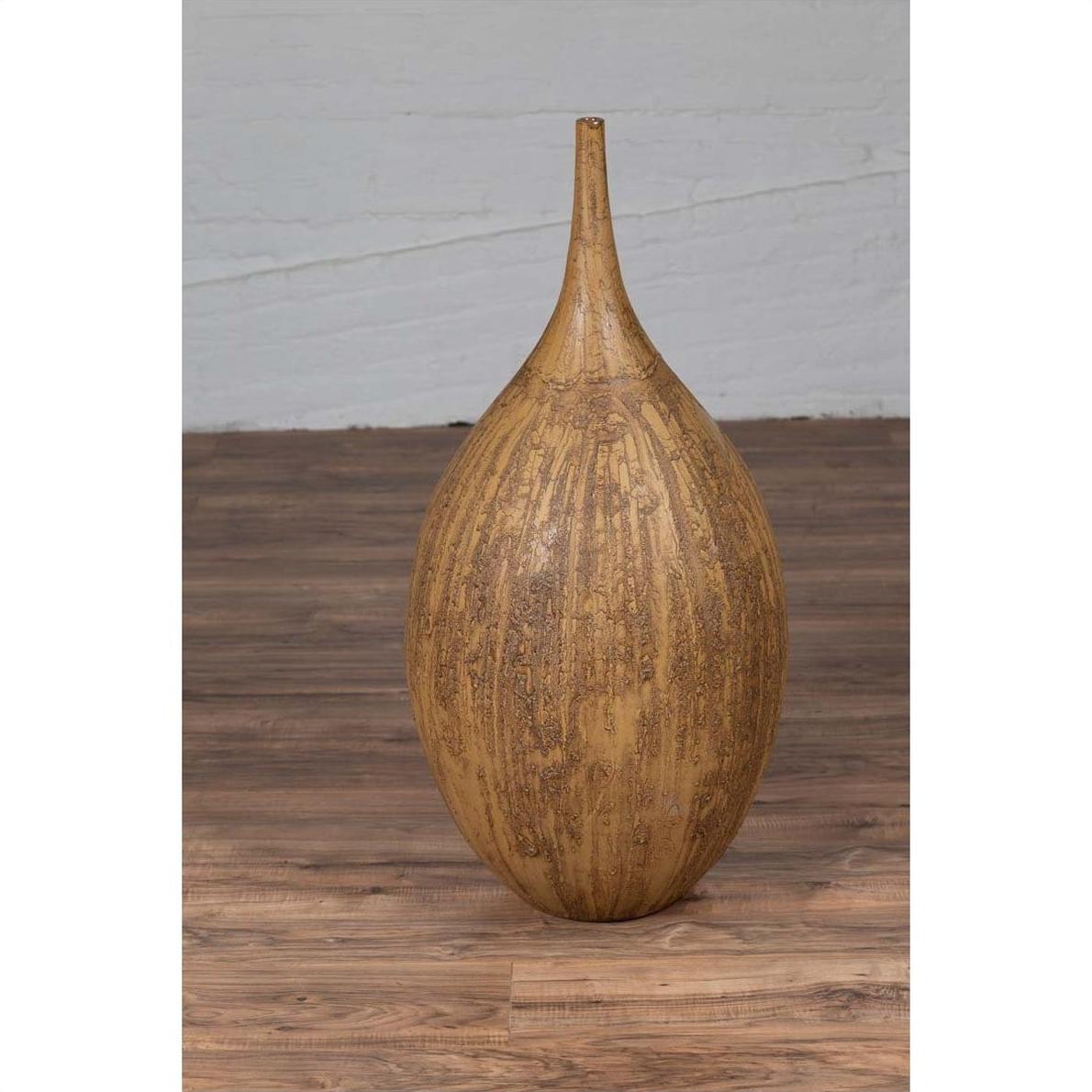 Mustard glazed ceramic vase with tapered spout on wooden floor beside a light gray wall indoors.
