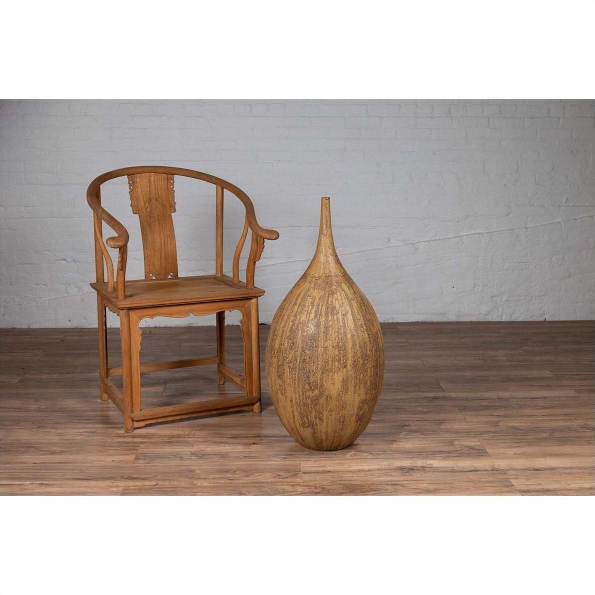 Wooden chair and mustard-glazed Thai ceramic vase on wood floor next to white brick wall indoors.