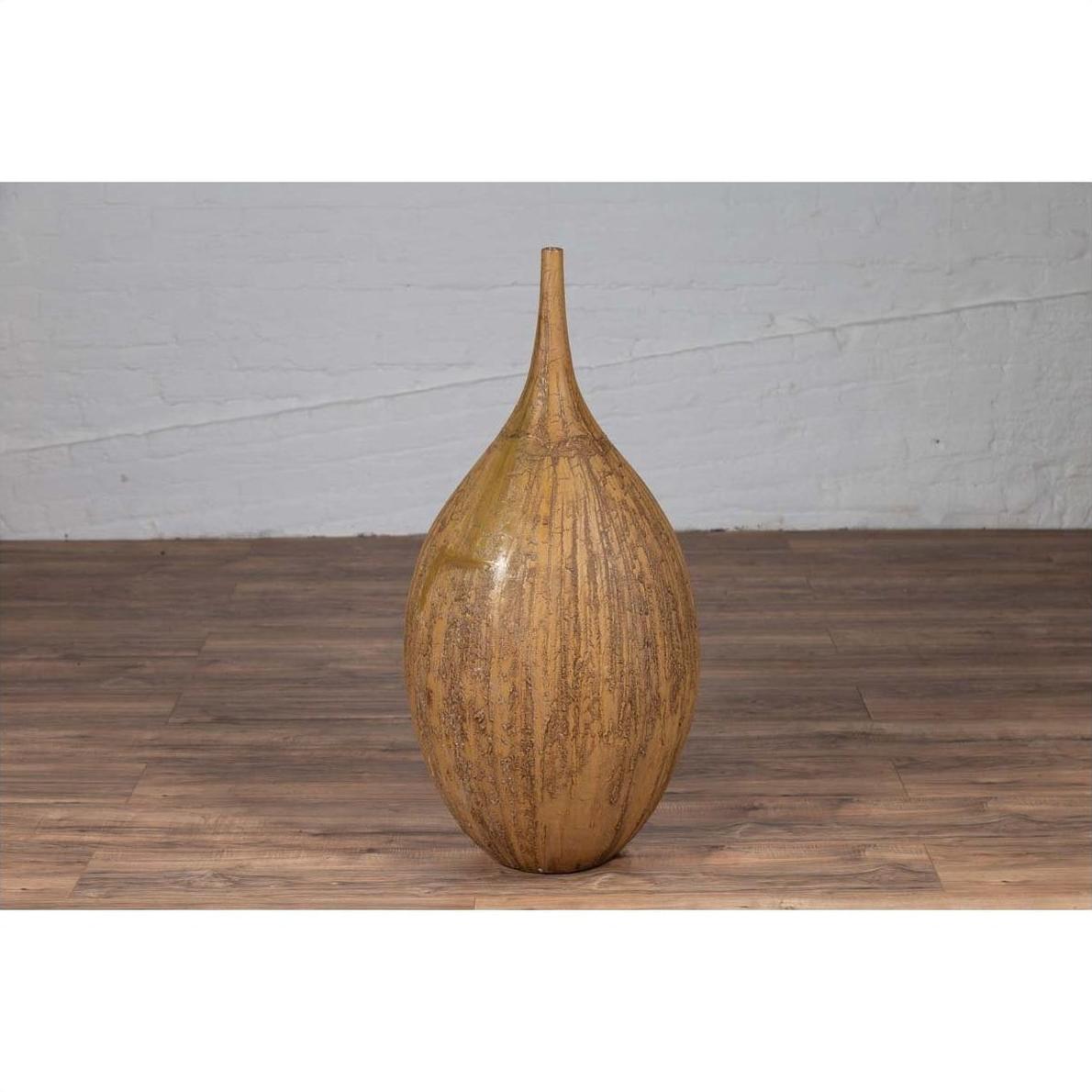 Mustard glazed ceramic vase with tapered spout on wooden floor, white brick wall in background.