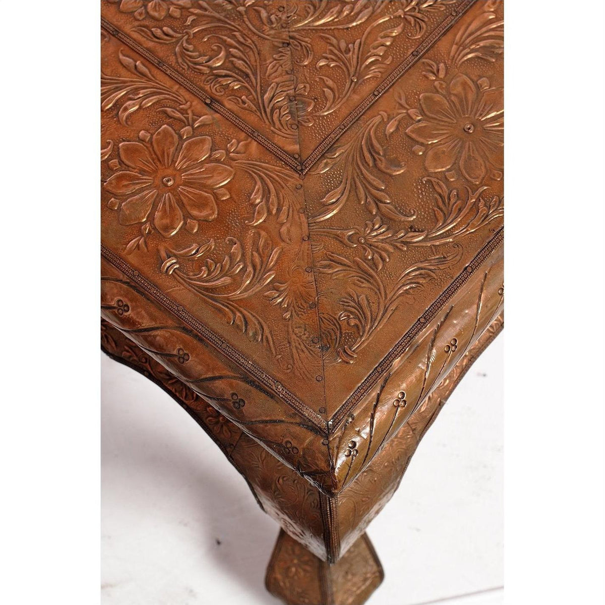 Close-up of copper coffee table corner with embossed floral and leaf patterns on the metal surface and edges.