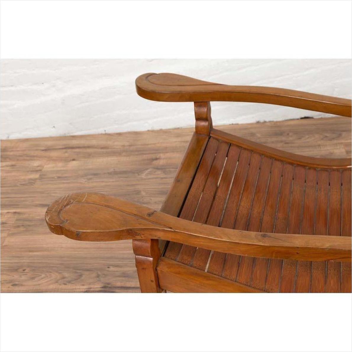 Brown wood lounge chair with curved seat and slats on a wooden floor next to a white wall indoors.