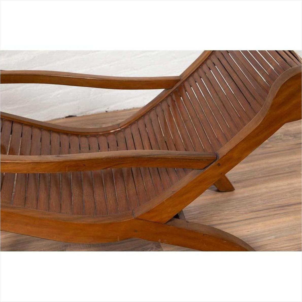 Wooden lounge chair with curved seat and slats, on wooden floor against a white wall indoors.