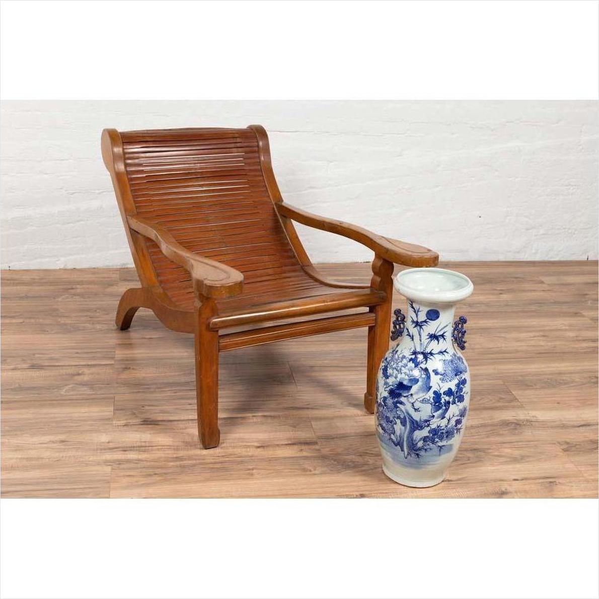 Brown wood Indonesian lounge chair with curved seat on wooden floor next to tall ceramic vase indoors.