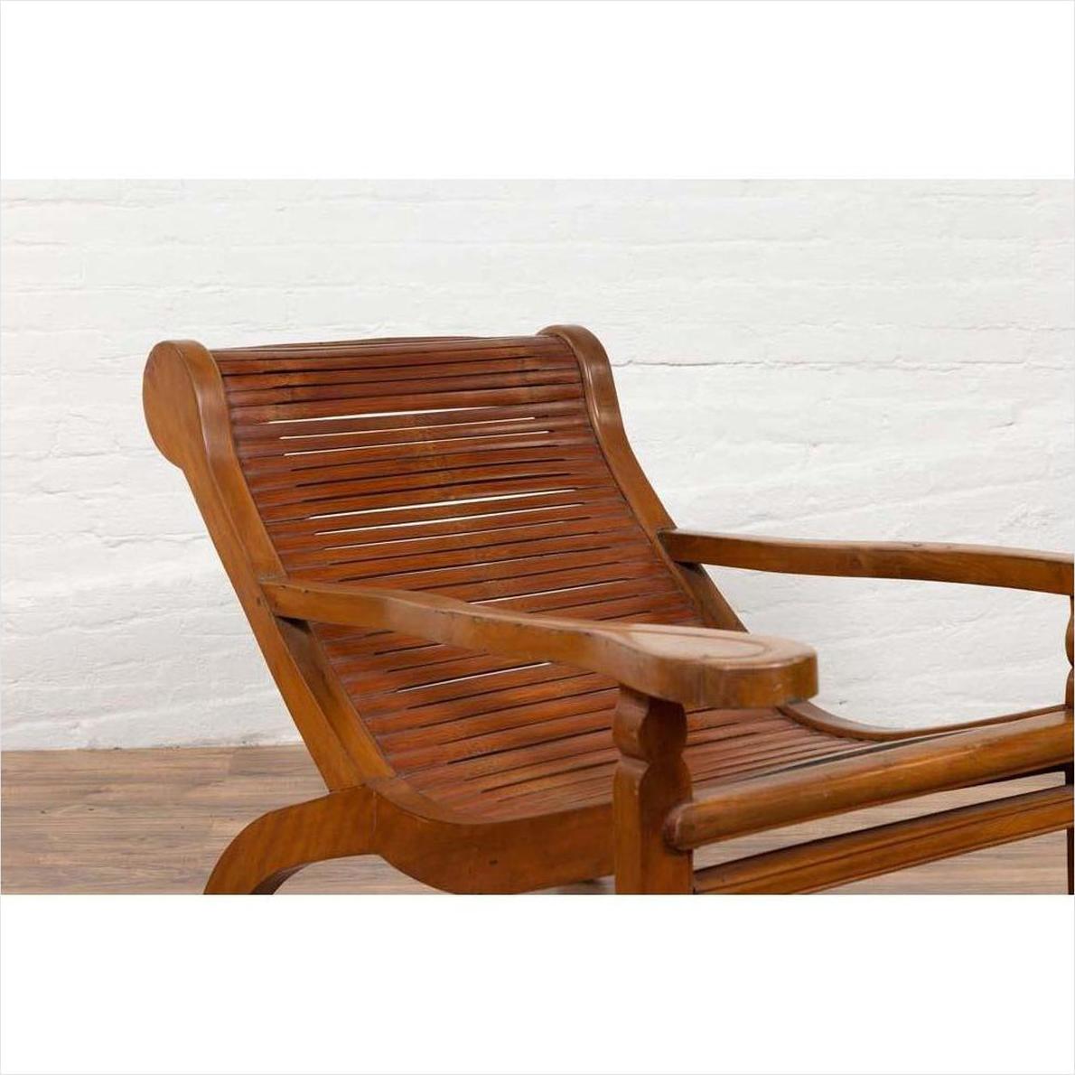 Brown wood lounge chair with curved seat and slats, placed indoors by a white brick wall on a wooden floor.