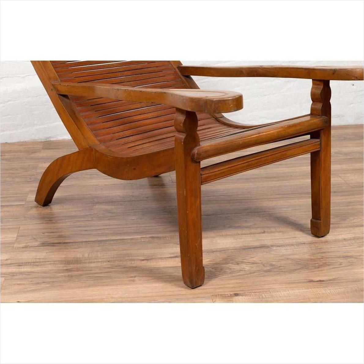 Wooden Dutch Colonial Indonesian lounge chair with curved seat and slats on a wooden floor near a white brick wall.