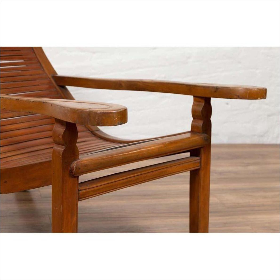 Close-up of a brown wooden lounge chair with curving seat and slats on a wooden floor against a white wall indoors.