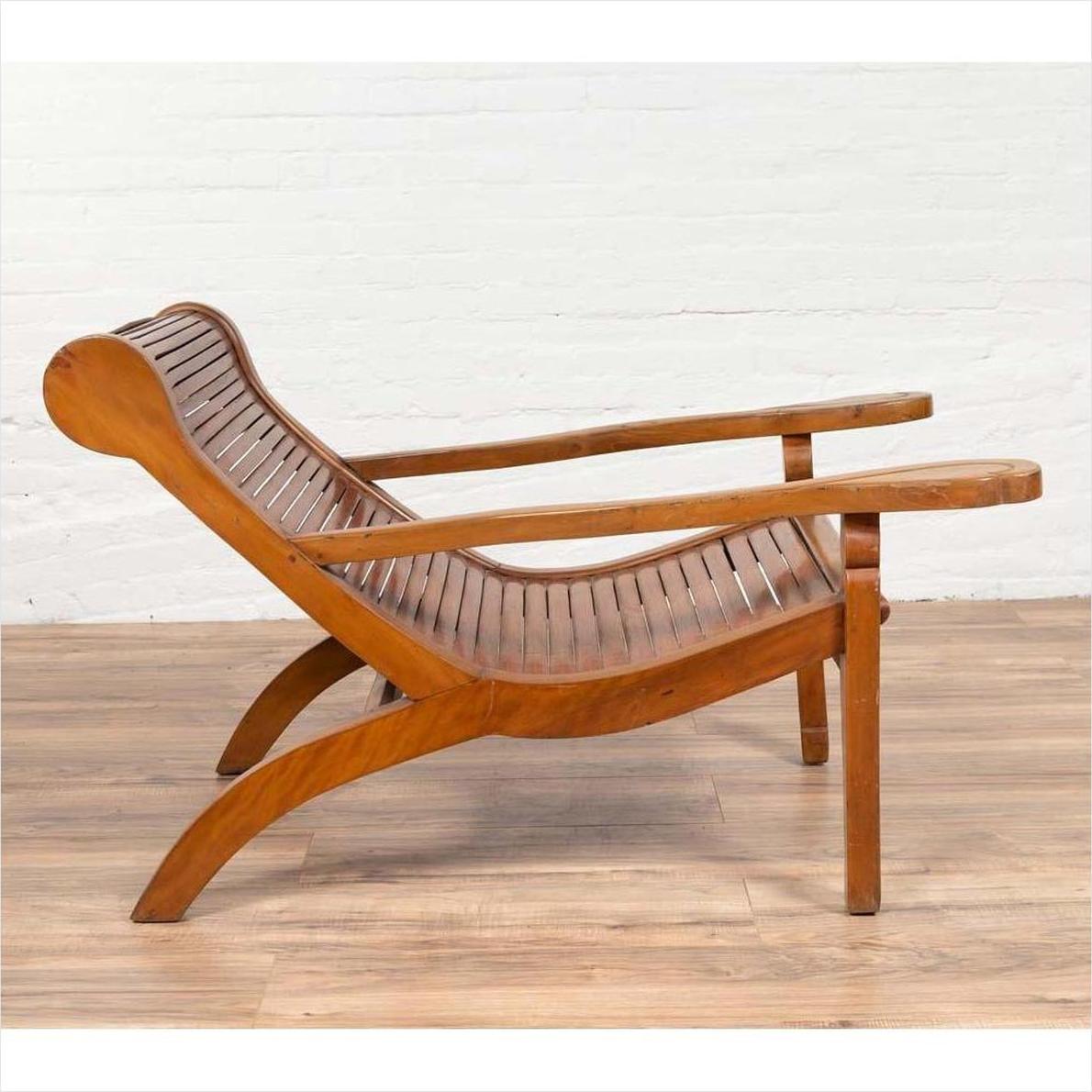 Brown wood plantation lounge chair with curved slatted seat, side view, on wooden floor next to white brick wall indoors.