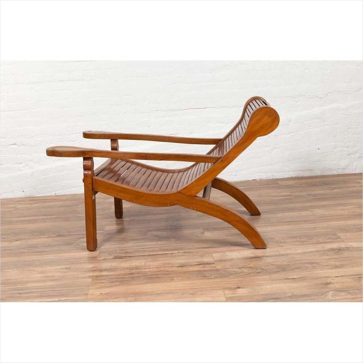 Brown wood lounge chair with curving seat and slats, on wooden floor next to white brick wall indoors.