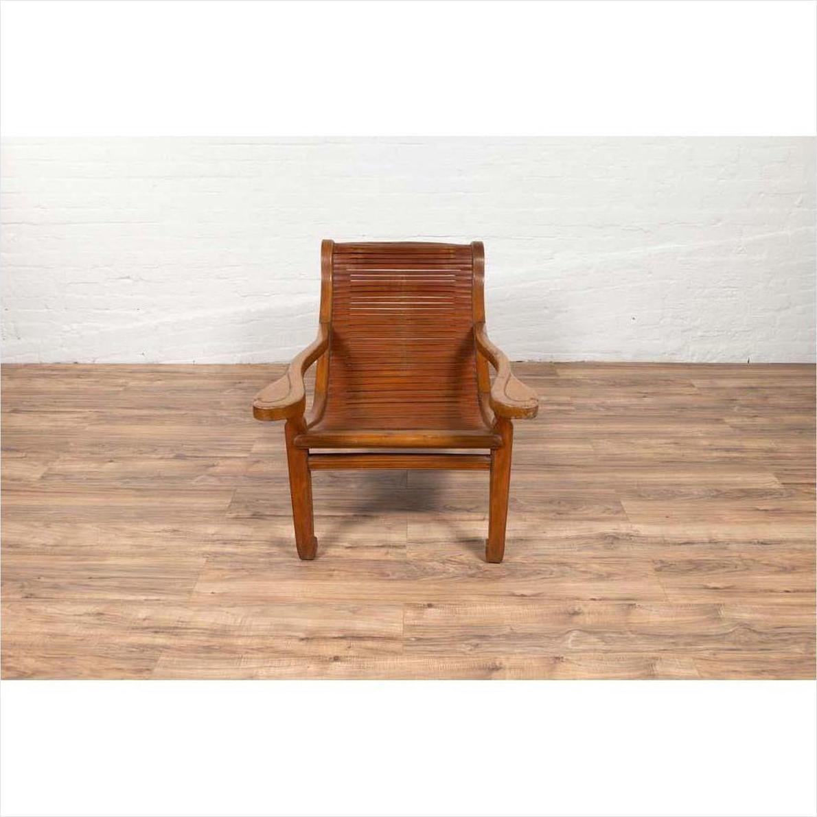 Wooden Dutch Colonial Indonesian lounge chair with curved seat and slats on a wooden floor by a white brick wall.