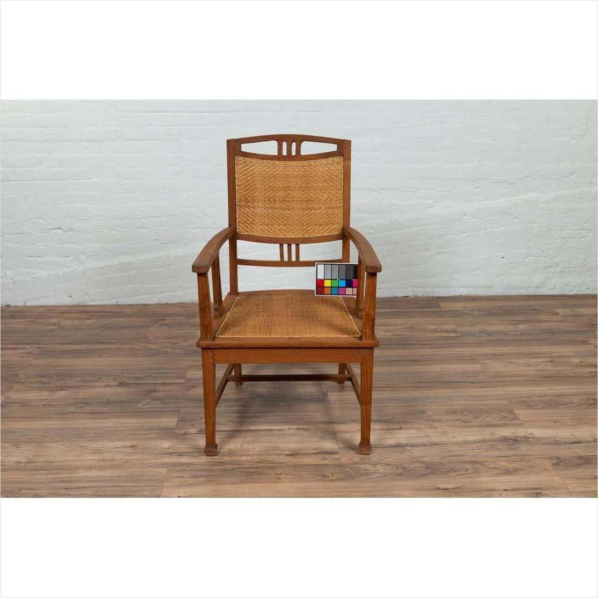 Brown teak and rattan Dutch Colonial Javanese armchair with triglyph motifs on wooden floor by white brick wall.