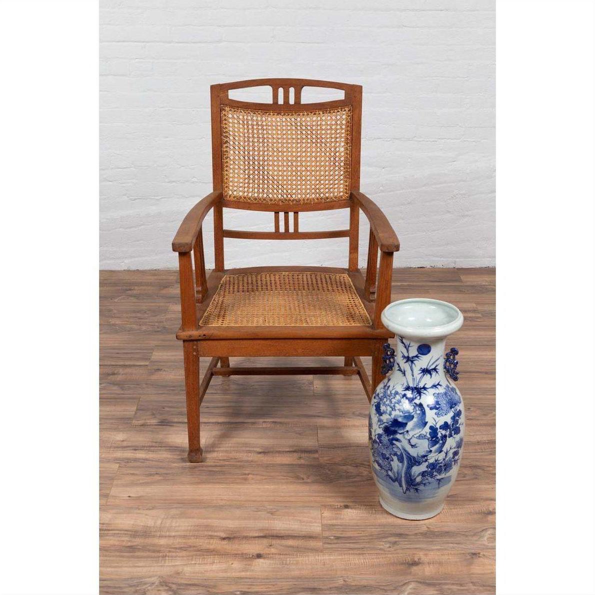 Brown teak armchair with rattan seat beside tall blue and white ceramic vase on wood floor, side view.