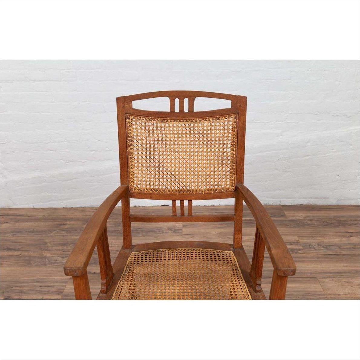 Brown teak and rattan Dutch Colonial Javanese armchair with triglyph motifs on wooden floor by white brick wall.