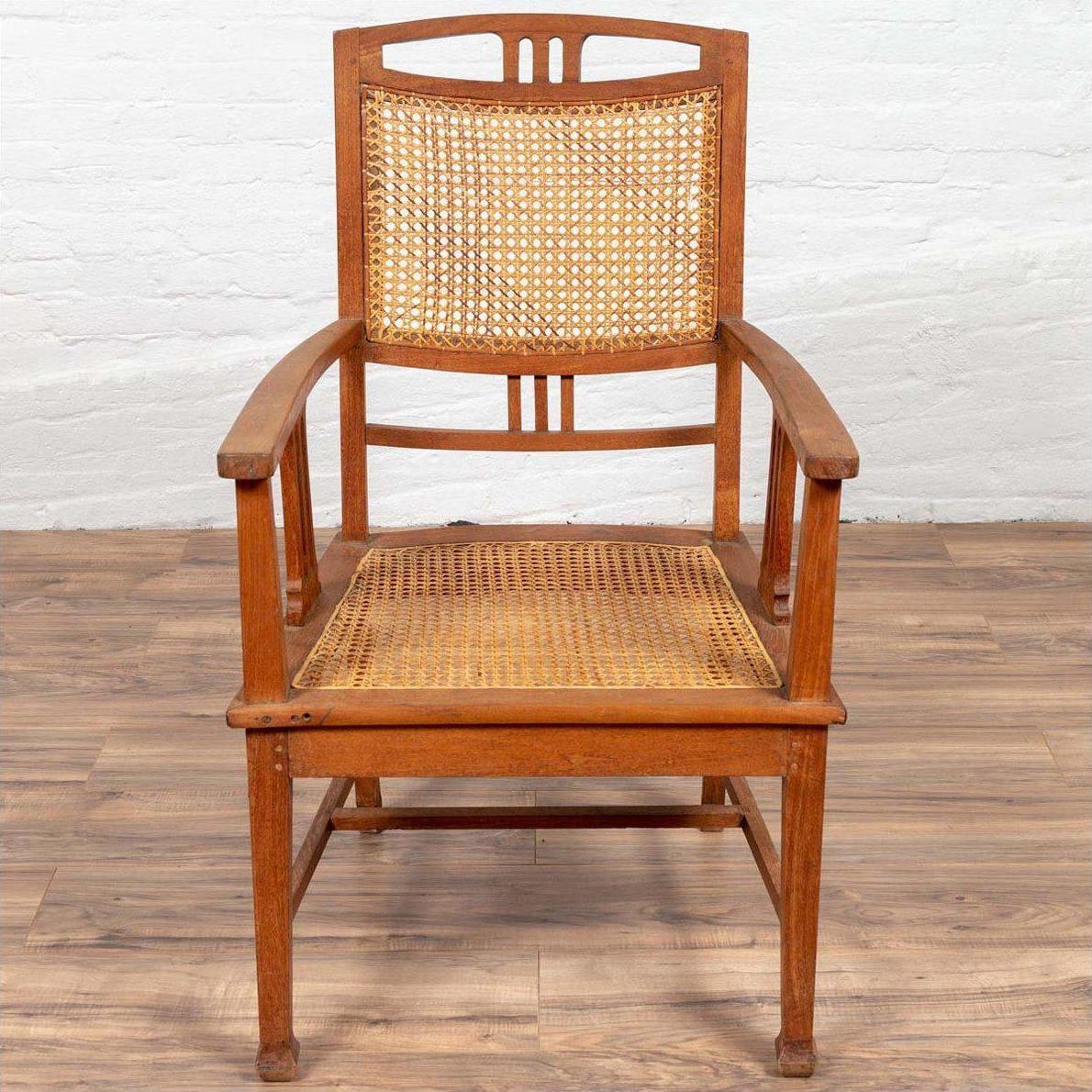 Brown teak and rattan armchair with triglyph motifs on wooden floor against a white brick wall.