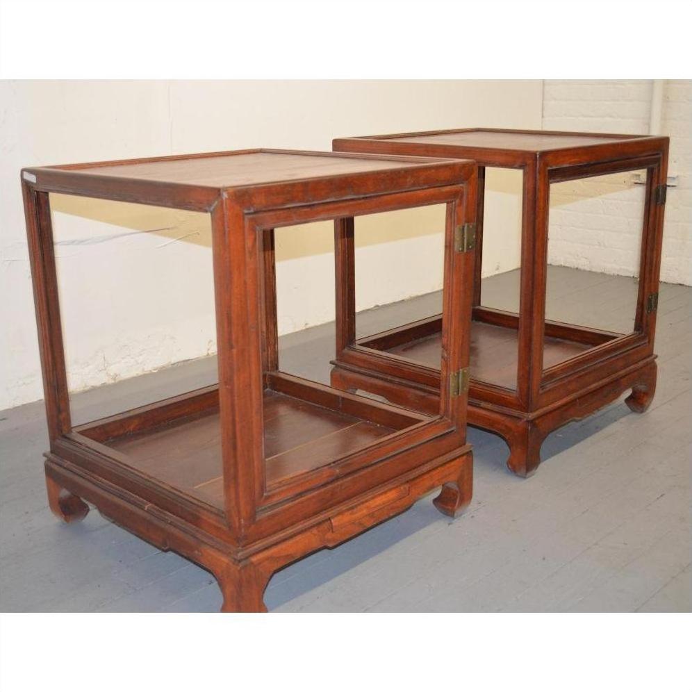 Brown Elmwood side tables with glass sides and hinged doors, positioned side by side on a gray floor against a white wall.