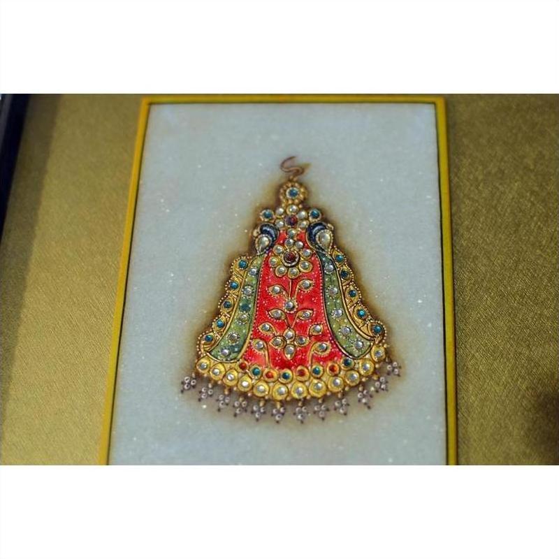 Close-up of framed Indian artwork with red, green, and blue enamel, gemstones, pearls on white fabric.