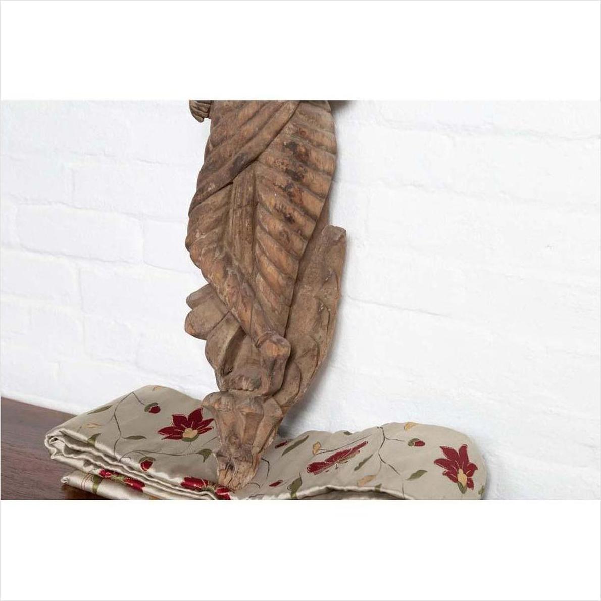 Brown wooden Indian deity statue on floral-embroidered fabric, white brick wall background, indoor setting.