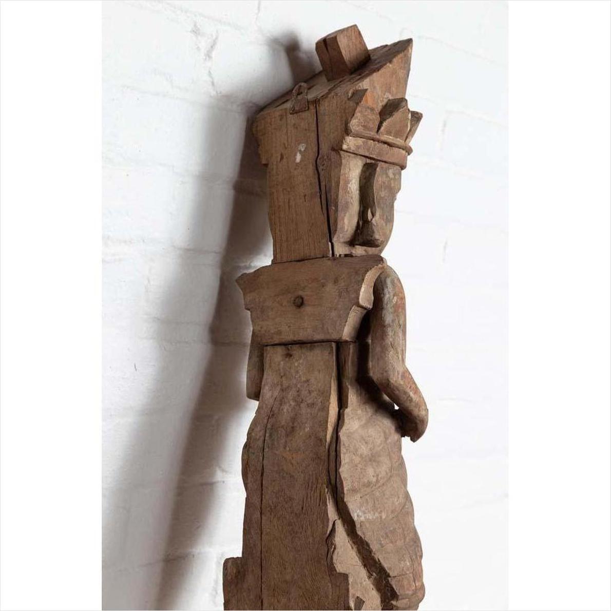 Brown hand-carved wooden statue of a Hindu deity from Gujarat, set against a white brick wall indoors.
