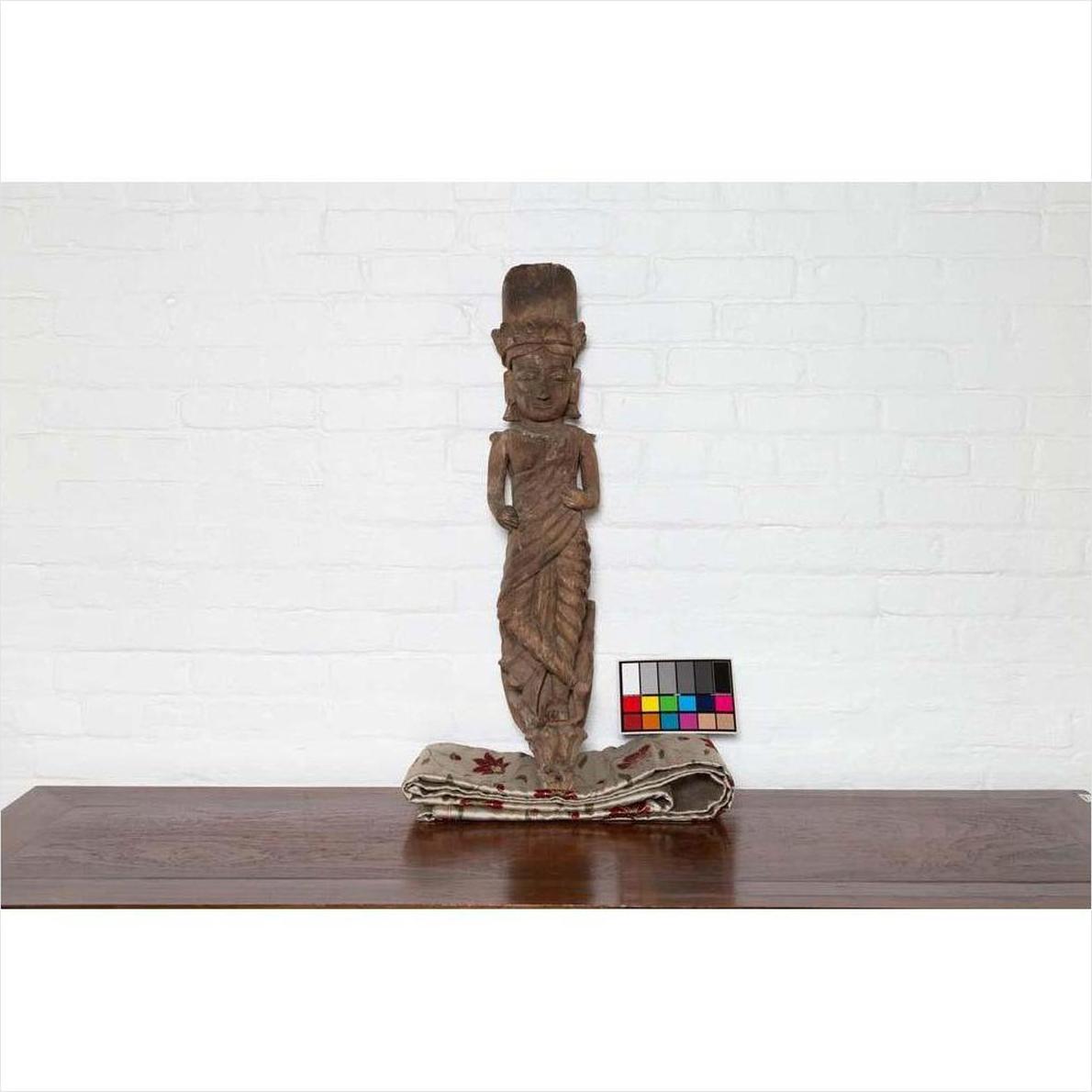Brown wood carved statue on folded textile next to color checker, white brick wall background.