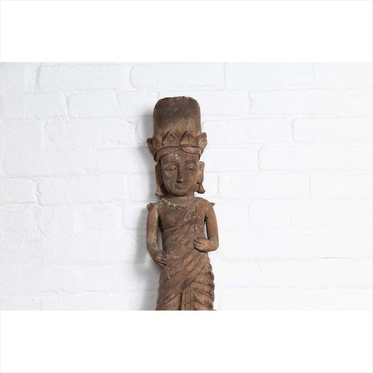 Brown wood hand-carved Hindu deity statue from Gujarat, indoors against a white brick wall.