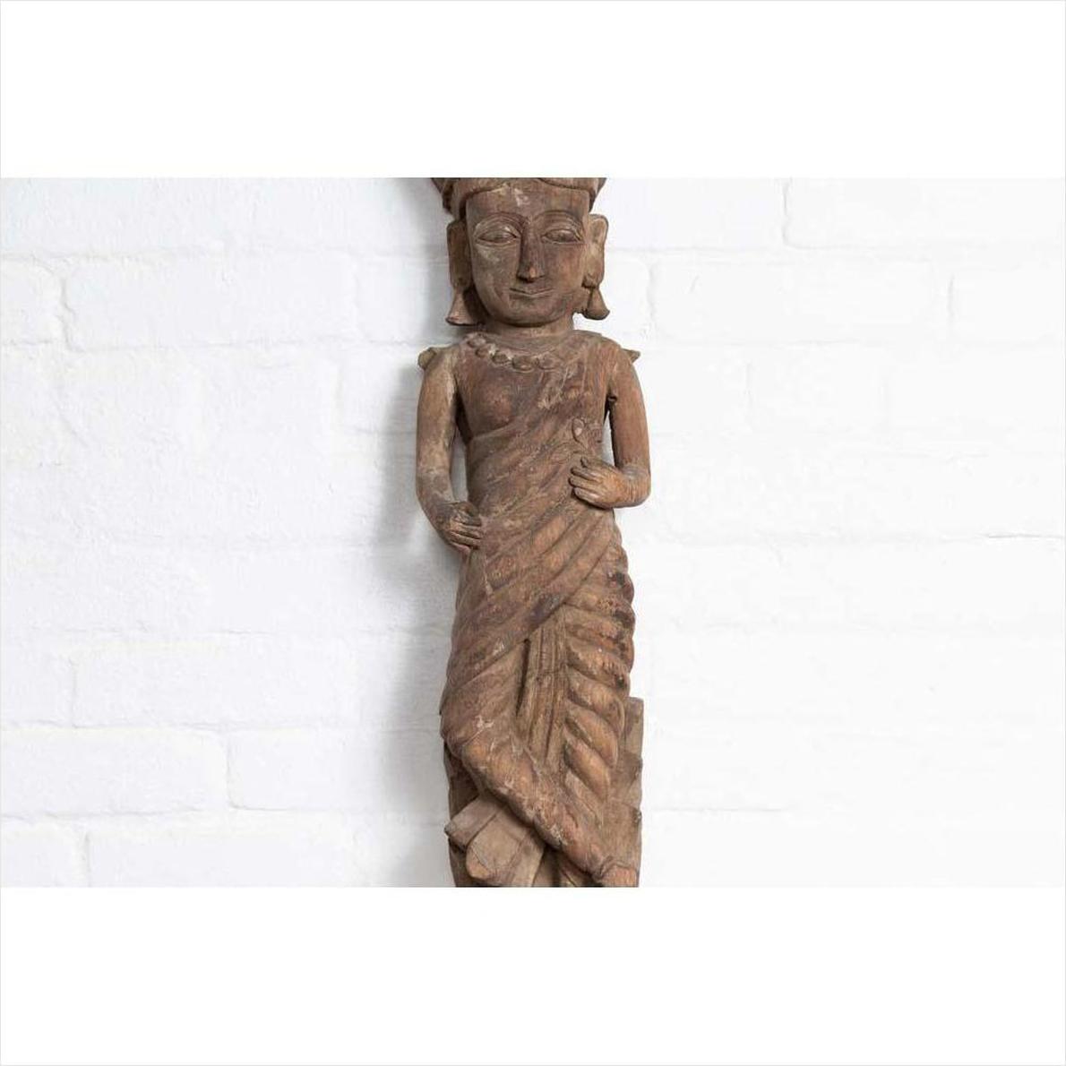 Brown wood hand carved Hindu deity statue from Gujarat, displayed indoors against a white brick wall.